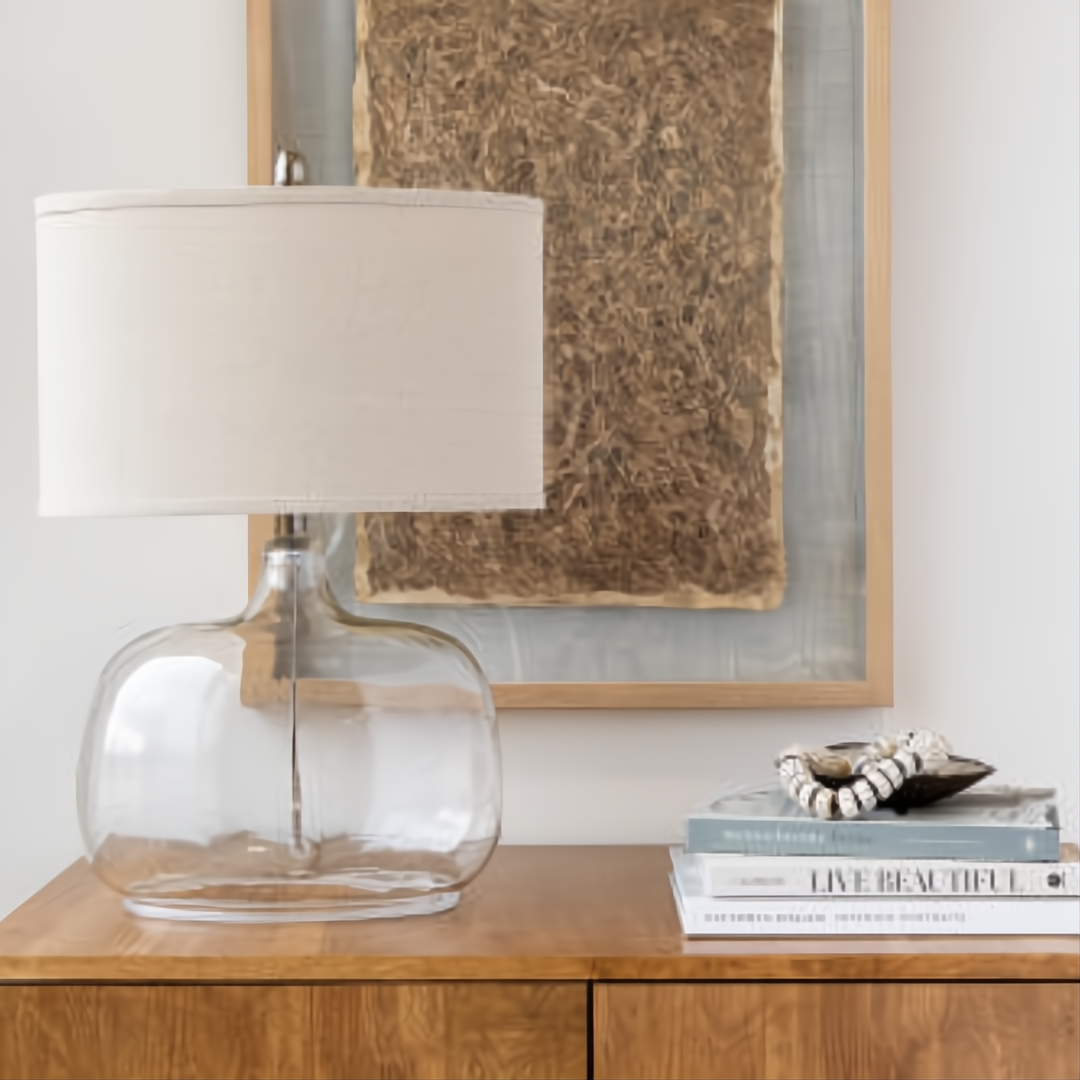 Surya Bentley accent table lamp with clear glass base, natural fabric shade, and metal accents on a wooden surface.