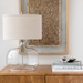 Surya Bentley accent table lamp with clear glass base, natural fabric shade, and metal accents on a wooden surface.
