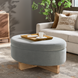Oval light green upholstered storage ottoman with a natural wood base, topped with a gold tray and coffee cup.