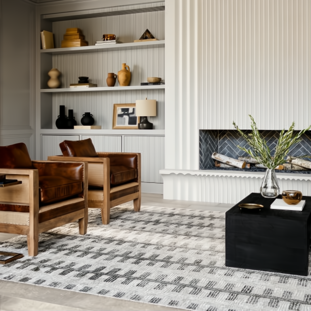 Modern living room with a light gray patterned rug, two brown leather and wood chairs, and a white fireplace.