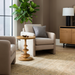Round Surya Adour end table with a brown oak base and refined wood top, next to a beige accent chair.