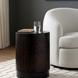 Round dark brown end table with hammered texture, black base, and a glass of water, next to a white armchair.