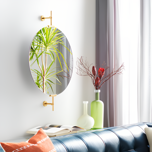 Modern oval mirror with gold-finished wall mounts, reflecting a plant, above a blue sofa and decorative vases.