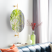 Modern oval mirror with gold-finished wall mounts, reflecting a plant, above a blue sofa and decorative vases.