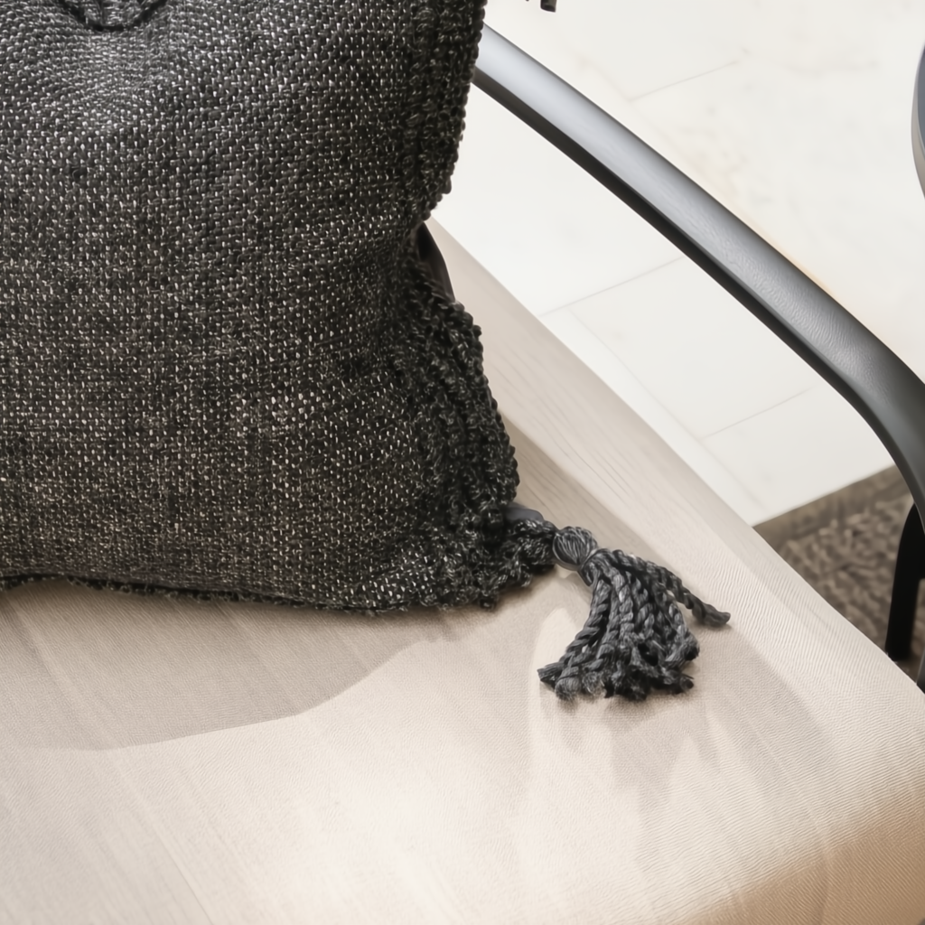 Close-up of a black textured throw pillow with a tassel on a beige outdoor loveseat with a black aluminum frame.