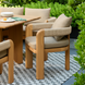 Taupe Sunbrella outdoor dining chair with a teak wood base and woven armrests on a checkered rug.