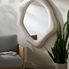 White lacquered concrete accent mirror with a specialty shape, mounted above a chair and next to a plant.