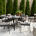 Black aluminum outdoor dining table with six striped chairs, set on a patio with brick wall and evergreens.