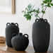 Three black ceramic decorative accent vases with handles, one holding green leaves, on a wooden surface.