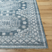 Surya Almeria low pile area rug with intricate geometric patterns in blue, gray, and cream, on a wood floor.