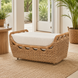 Light brown woven rattan outdoor ottoman with a cream cushion on a natural fiber rug.