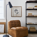 Brown leather modular chair with knife-edge design, black base, next to a black floor lamp and bookshelf.