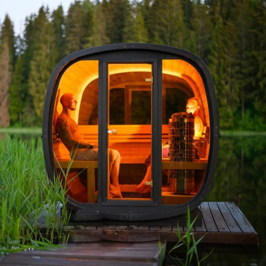 Luxury outdoor cedar barrel sauna with two people inside, glowing warmly by a lake at dusk.